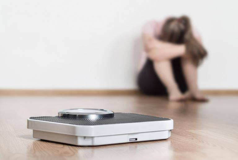 A scale on the floor with a woman sitting in the background with her head hung with failure.